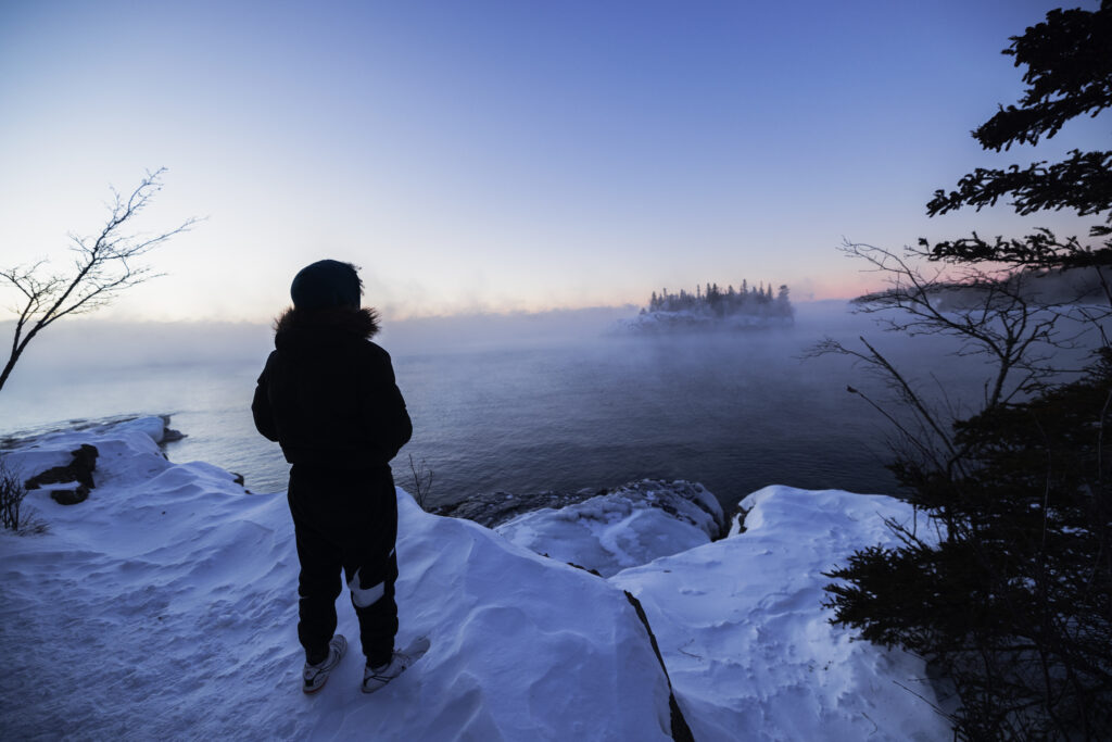 The silhouette of a person standing on a snowy rock looking out across a lake with steam rising into the cold air.