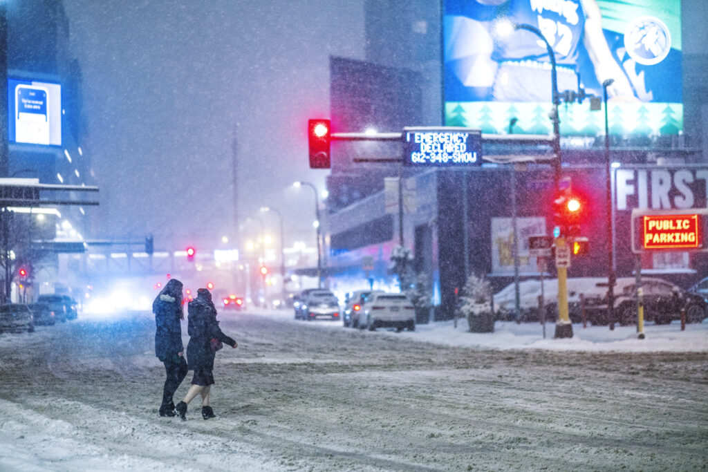 Two people bundled up cross a snowy street in downtown Minneapolis near First Avenue.