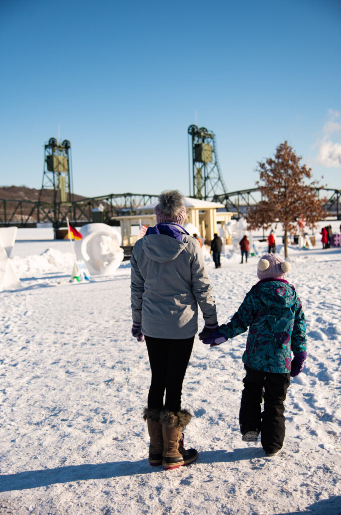 An adult and child stand holding hands looking at ice sculptures near a river with a lift bridge. The one sculpture we can see appears to be a round bust of a woman.
