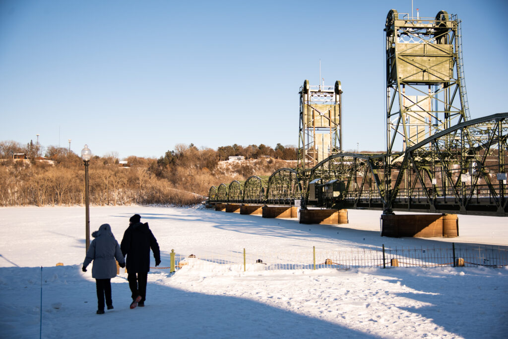 Two people walk hand-in-hand along a frozen river near a lift bridge.