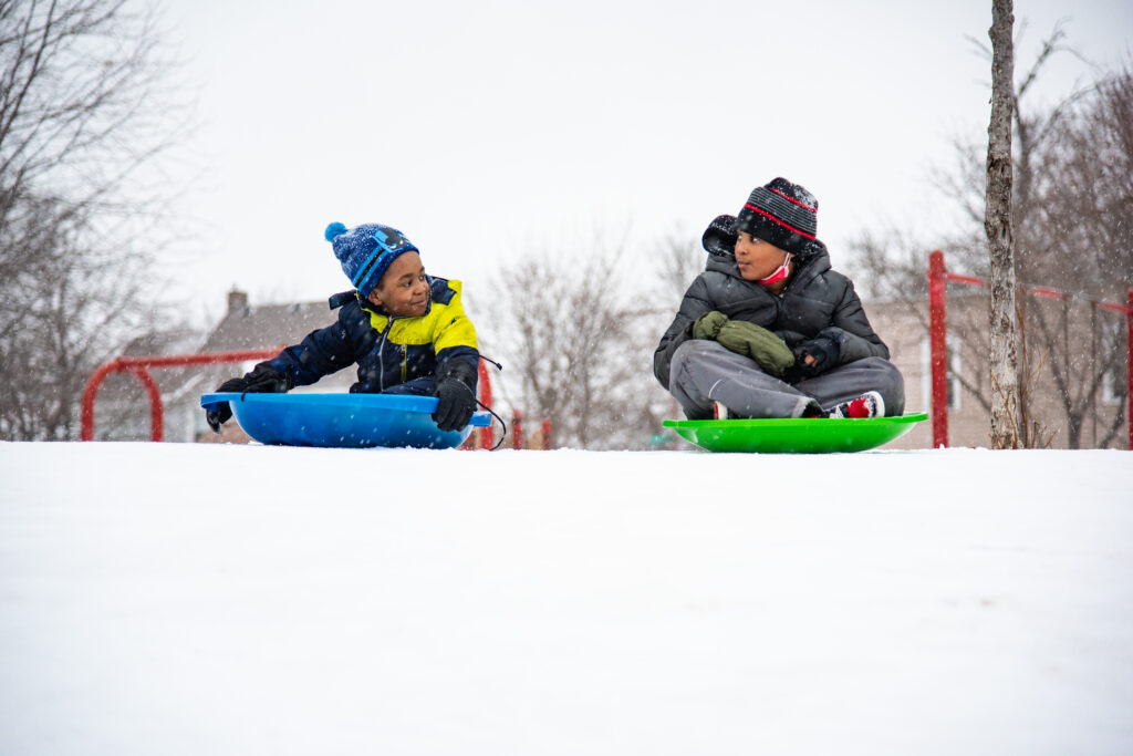 Two children, one in a blue hat and yellow and black jacket on a blue sled and one in a gray jacket on a green sled, look at each other before going down the hill.
