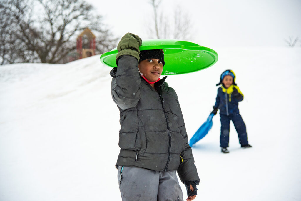 A girl in all gray balances a lime-green disc sled on her head while a young boy watches behind her.