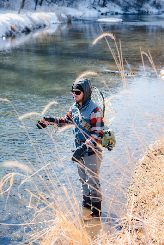 A person in snow boots, a plaid shirt, and denim vest stands fishing in a cold stream.