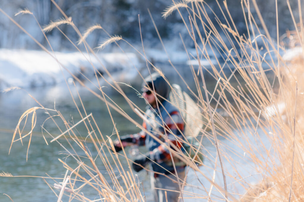 A person in snow boots, a plaid shirt, and denim vest stands fishing in a cold stream.