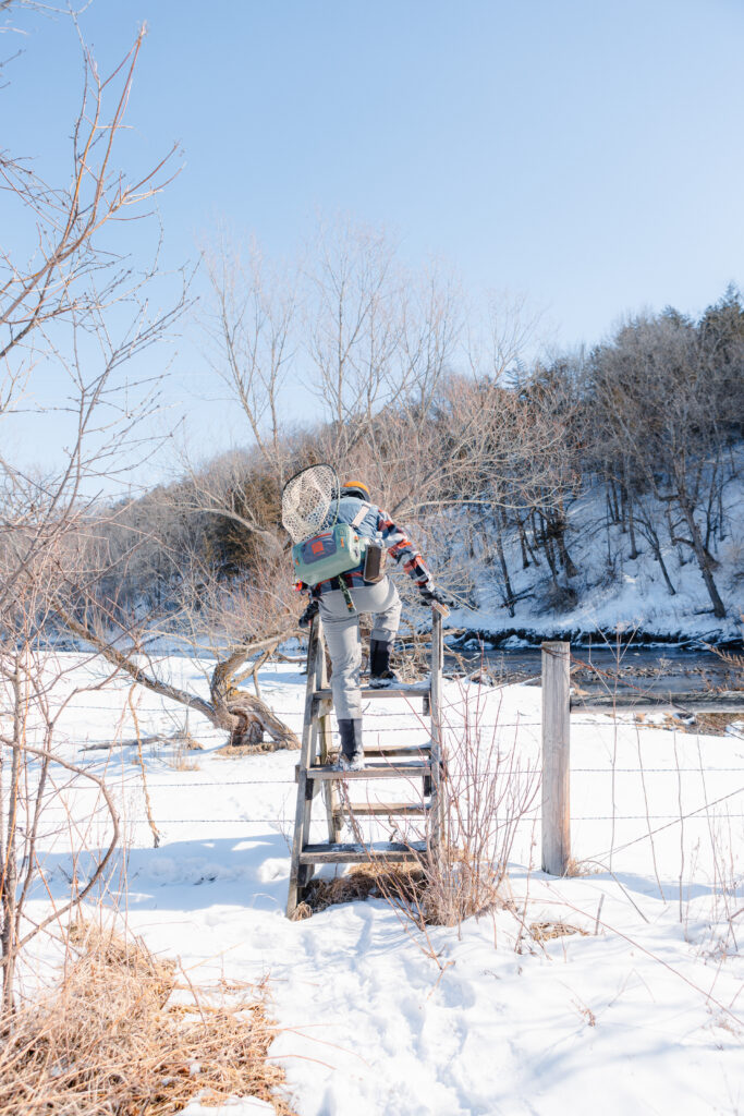 A person in snow boots, a plaid shirt, and denim vest climbs up on a ladder to get over a barbed-wire fence toward a river.