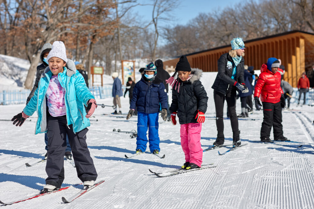 A large group of kids on skis, all in colorful coats, hats, and snowpants, cross country ski toward the camera on a snowy trail.