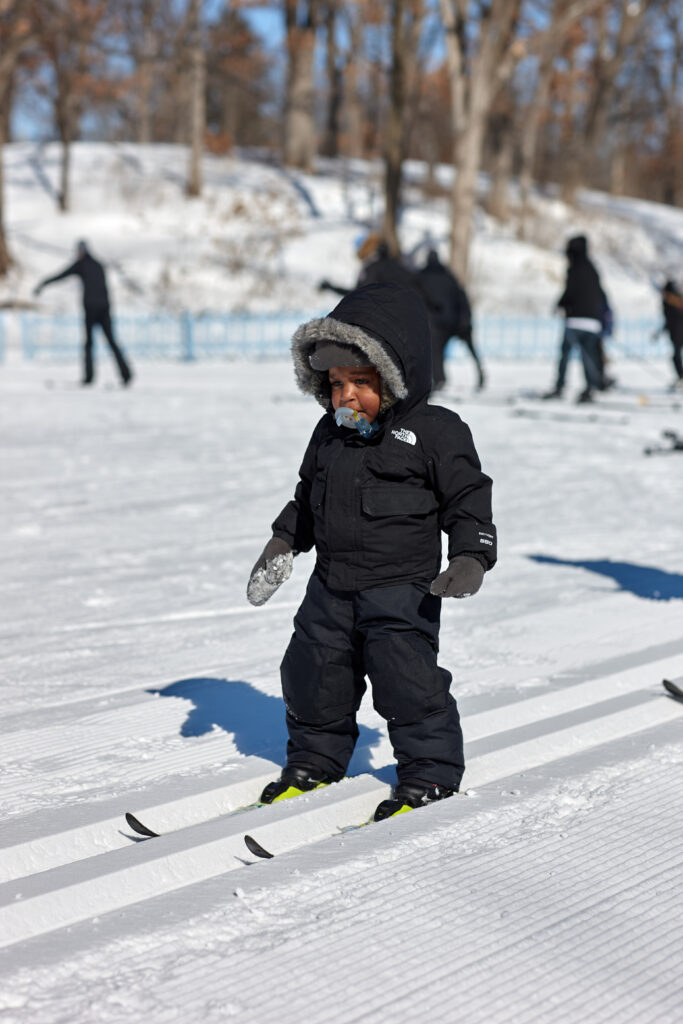 A young boy in a black snowsuit balances on two skis with a pacifier in his mouth.