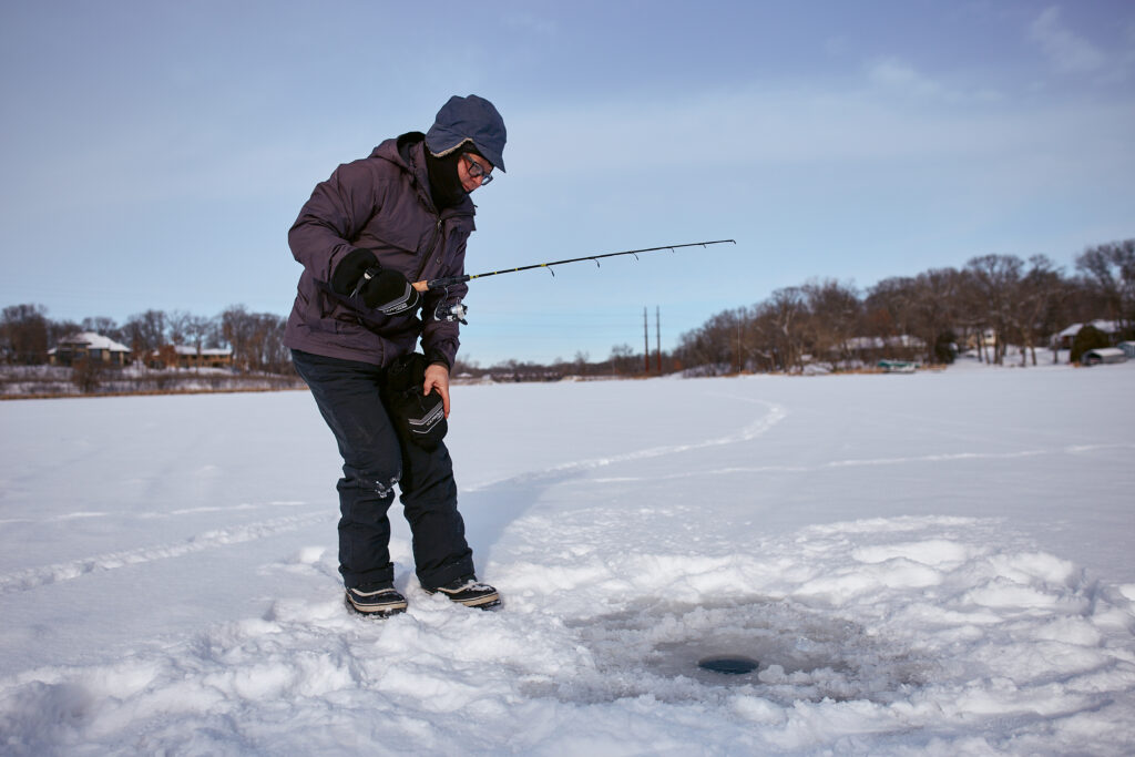A man in all dark clothing leans over an ice fishing hole with a rod.