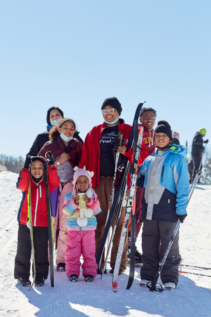 A family of seven in colorful coats, snowpants, hats, and mittens, stand and smile at the camera, all of them holding skis.