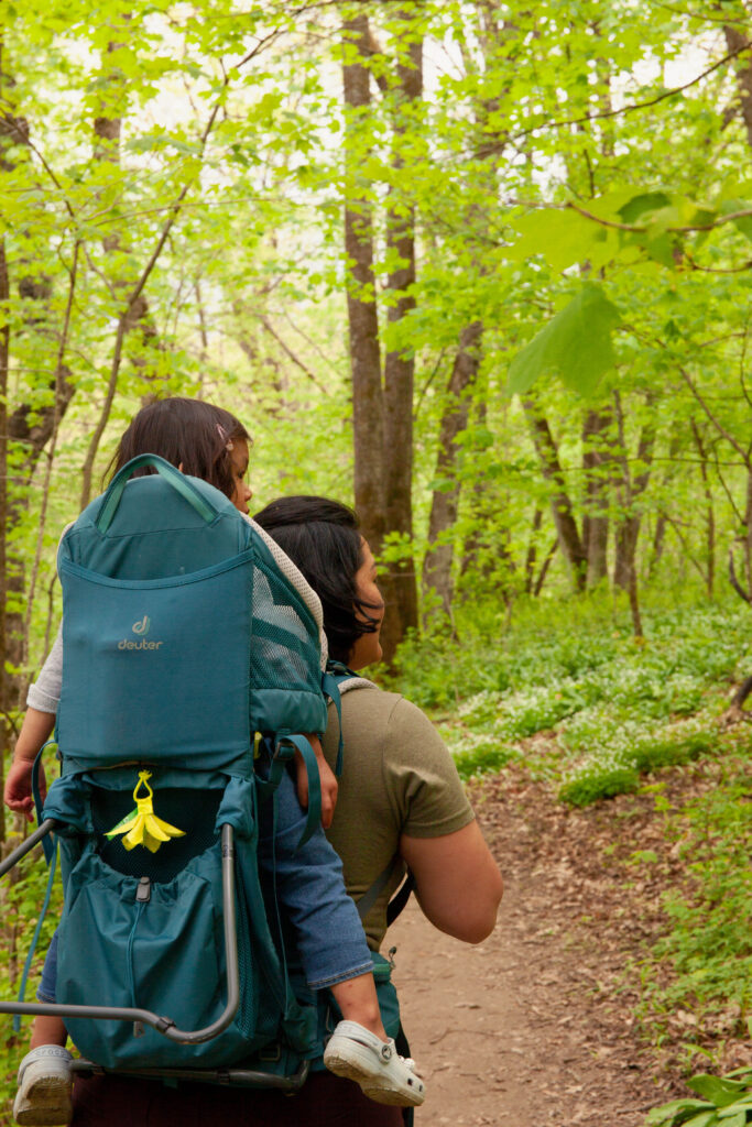A woman stands on a path in a green forest, holding a young girl on her back in a blue backpack.