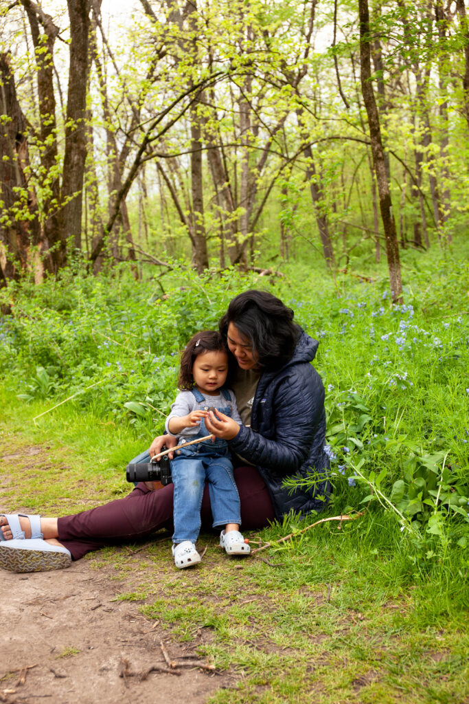 A young girl rests on her mothers lap as they look at flowers together on a forest floor.