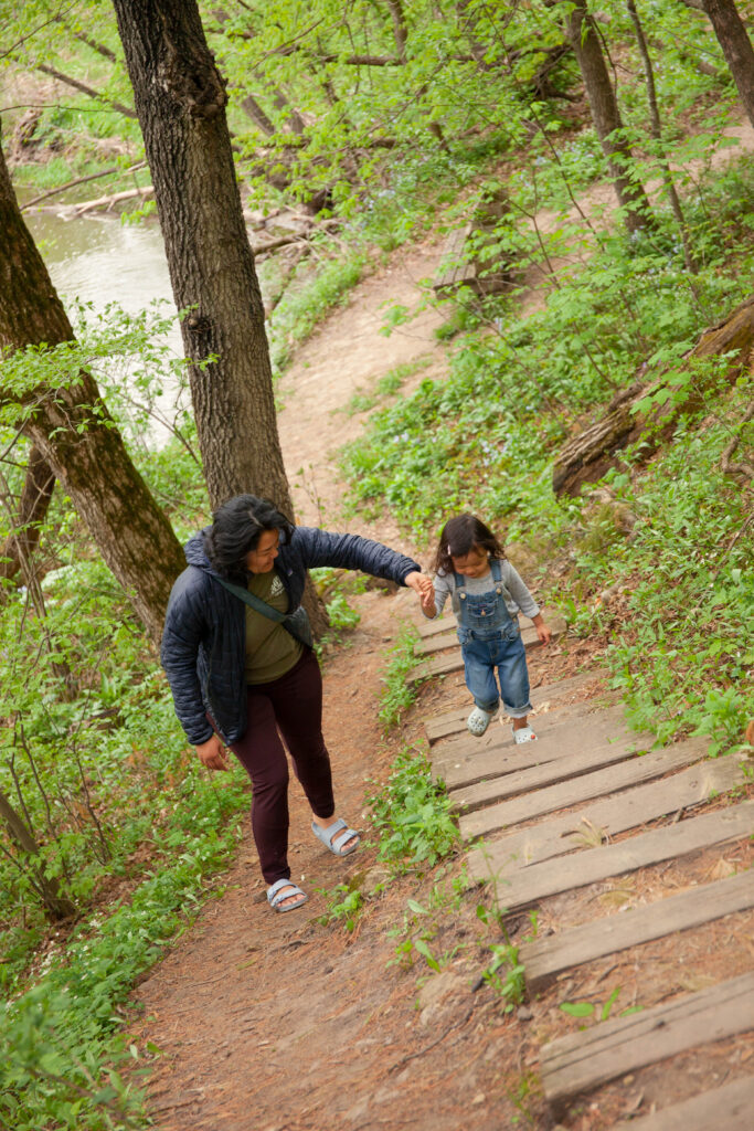 A woman holds the hand of a young girl in blue overalls as she walks up steps on a forest path.