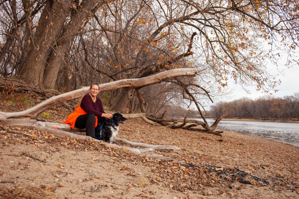A woman in a red shirt with a bright orange jacket tied around her waist sits on a sandy bank near a river with her black-and-white dog sitting beside her.
