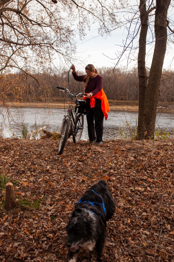 A woman in a red shirt with a bright orange jacket tied around her waist walks with her bike along a path full of fallen leaves.
