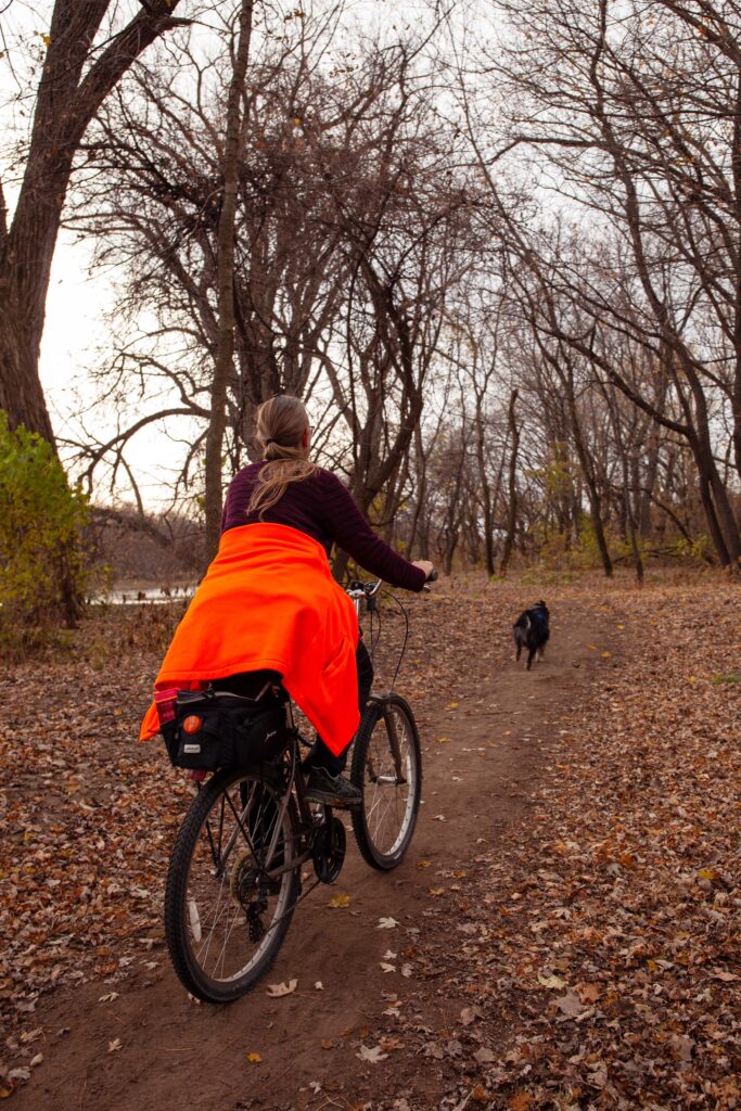 A woman in a red shirt with a bright orange jacket tied around her waist rides her bike down a trail full of fallen leaves while her black-and-white dog runs ahead of her.