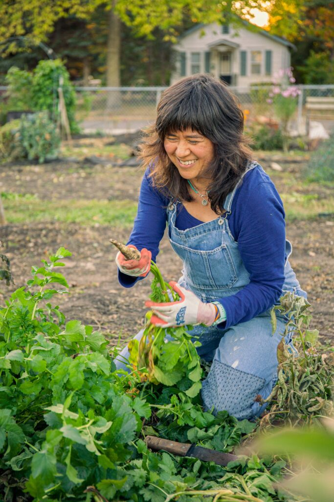 A woman with dark hair wearing a dark blue shirt and blue overalls kneels in a garden holding a freshly harvested carrot.