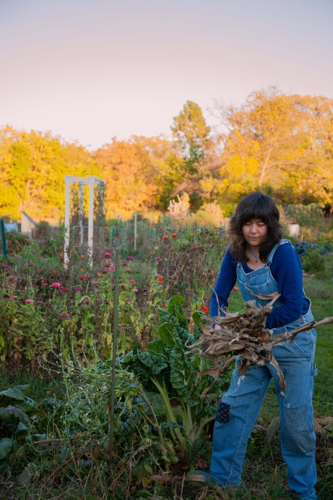 A woman with dark hair wearing a dark blue shirt and blue overalls bends collecting brush from a garden.