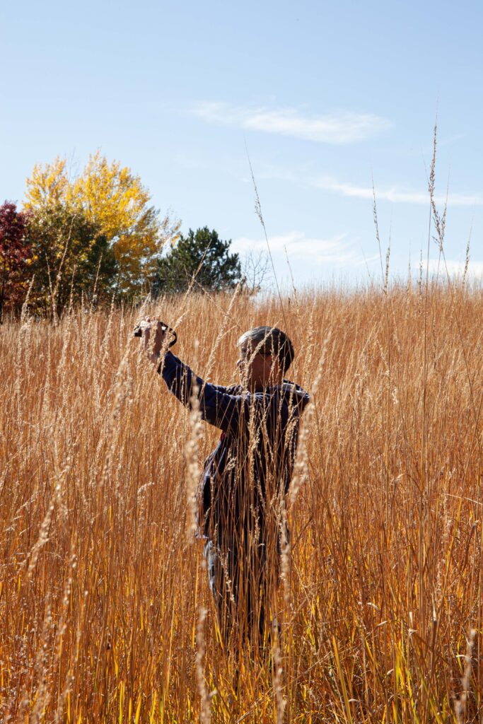 A man stands among golden tall grass and holds his camera high, taking a picture with a camera.