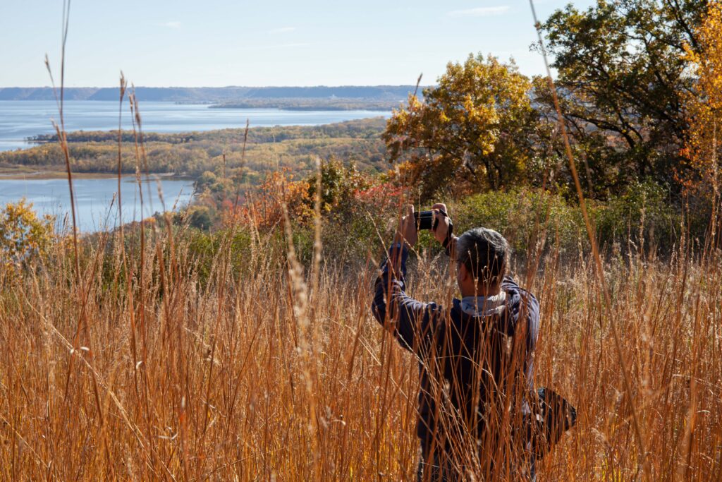 A man stands among golden tall grass and holds his camera high, taking a picture of a river.