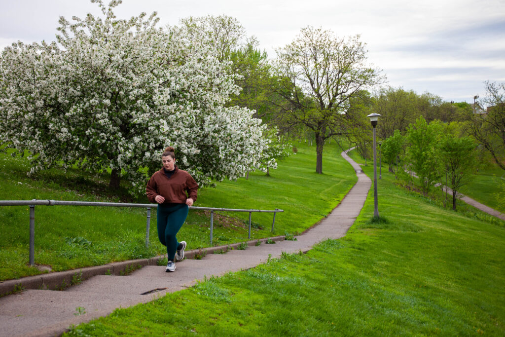 A woman in blue leggings and a red running jacket jogs down an asphalt path surrounded by flowering white trees.