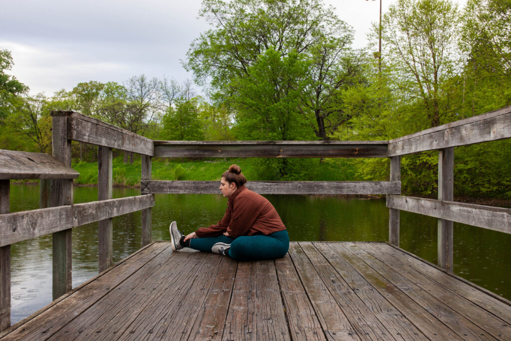 A woman in blue leggings and a red running jacket stretches while sitting on a wooden structure overlooking a lake.